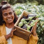 Joyful middle aged ethnic female farmer in casual clothes smiling and carrying wooden box with heap of fresh organic eggplants while working on plantation on sunny day